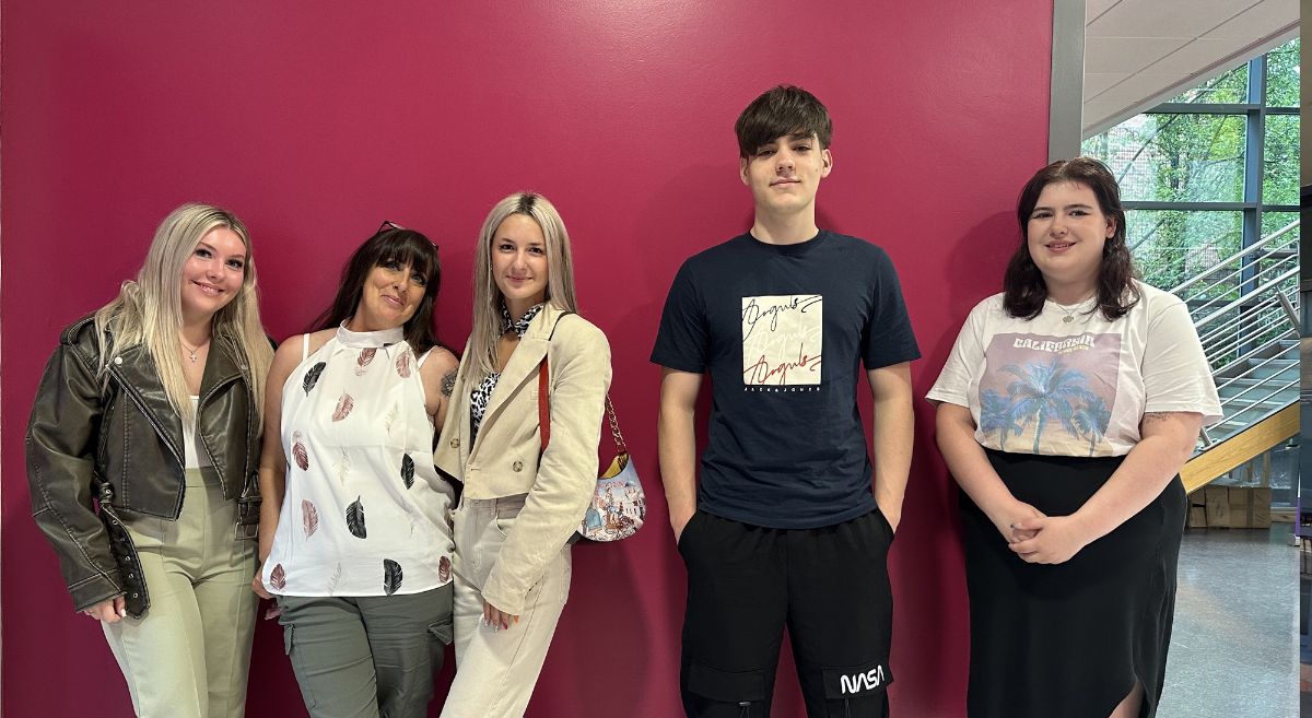 Five students, casually dressed, smiling to camera and pictured in front of a dark pink wall with staircase and window in background.